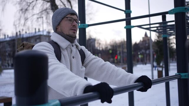 Determined man wearing a warm winter jacket, beanie, and gloves performs dips on parallel bars in a snow-covered park, demonstrating dedication to fitness despite the cold weather
