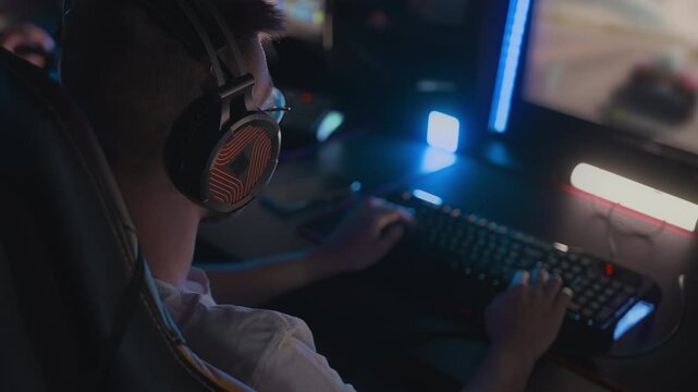 Gamer in glasses plays arcade racing video game on PC at computer club. Focus on professional headset with RGB light. Modern gaming keyboard and monitor. Cyber sport tournament E-sport championship