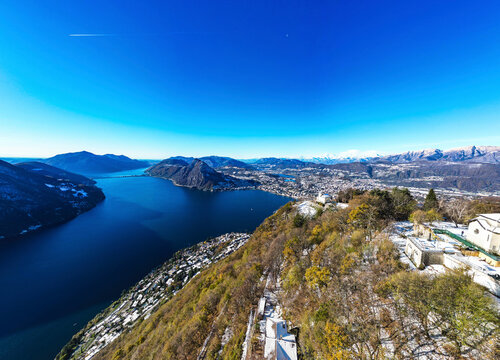Aerial view of beautiful Lake Lugano surrounded by majestic mountains and a picturesque town under a clear blue sky, Bre sopra Lugano, Switzerland.