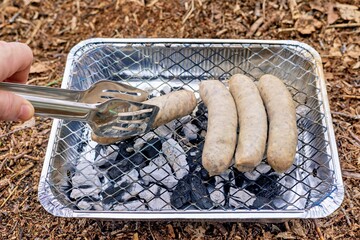 White sausages are fried on a disposable grill. His hand turns the sausage over with tongs.