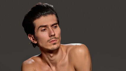 Fototapeta premium Man turns his head slightly, looking distant against dark background. His bare torso and subtle facial expression convey calm and reflective moment. Concept of male beauty and facial care procedures.
