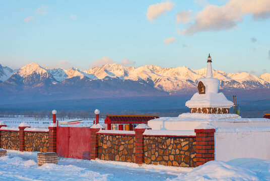 View of  Buddhist prayer complex with white stupa or suburgan and prayer drums against backdrop of snow-capped mountains of Eastern Sayan at winter sunset. Baikal region. Buryatia. Tunka valley