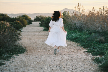 A woman in a flowing white dress runs down a gravel path, enjoying the serene sunset landscape. ...
