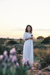 Serene woman in a flowing white dress stands in a field at sunset, holding wildflowers.  A peaceful and elegant image, perfect for blogs, websites, or design projects.