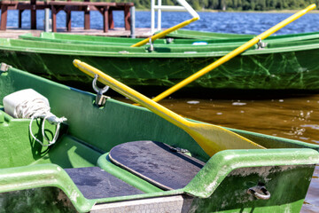 
On the shore of the lake, a green oar with a yellow blade is tied to a pier, which is in a green boat with a seat.