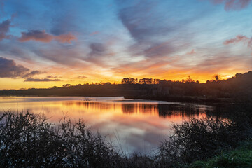 Lever de soleil aux teintes orangées se reflétant sur l'étang de l'Aber, presqu'île de Crozon. Quelques nuages se dessinent dans le ciel et se mirent dans l'eau paisible.