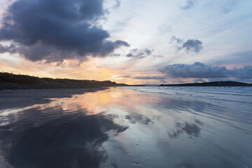 Sur la plage de l'Aber, un lever de soleil avec un ciel bleu clair et un jaune pâle, où quelques nuages gris se reflètent sur le sable mouillé, créant une ambiance douce et apaisante.