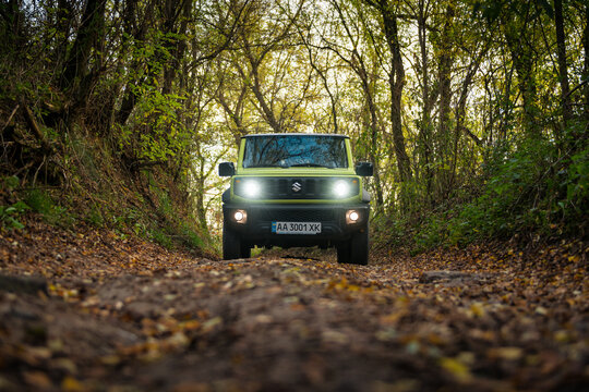 Lime green Suzuki Jimny in autumn forest. Front view of off-road mini SUV on dirt road in deciduous forest. Car with bright headlights driving towards camera. Kyiv region, Ukraine - October 25 2020.