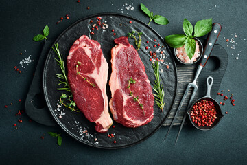 Two raw rib eye beef steaks with spices on a plate. Ready to cook. On a dark stone background. Top...