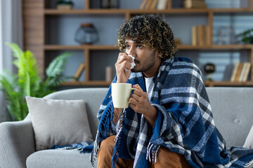 Sick man with a cold sits at home on the couch, covered with a blanket, and with a napkin in his hand. A man with a cold with a hot drink in his hands has a runny nose.
