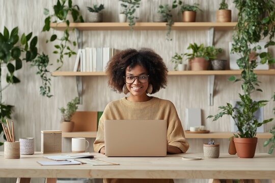 Smiling african american woman sitting at a desk with a laptop in a home office with shelves full of potted plants and books - Powered by Adobe