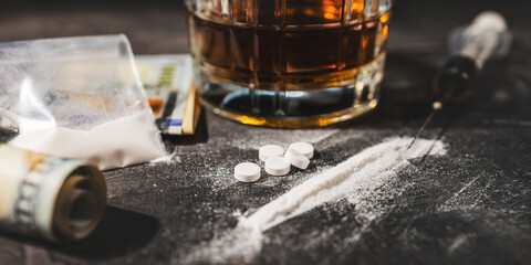 White drugs, narcotic pills, an alcoholic drink in a glass, a syringe with a dose of heroin, a line of cocaine powder and US dollar bills on a dark background. Concept of addiction and bad habits