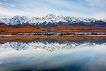 Obraz premium Scenic view of snow-covered mountains mirrored in calm lake surrounded by autumn forest in Altai.