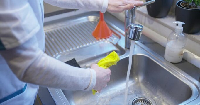Wringing a dishcloth under the kitchen faucet with water flowing, a hand turning off the tap, set in a home kitchen sink with hand soap nearby.