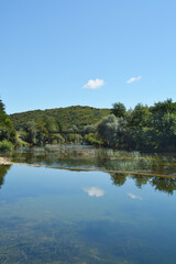 A peaceful scene of the Dobra River near Jaskovo, showcasing its clear waters reflecting the sky and surrounding greenery, a small dam, and an old bridge in the distance