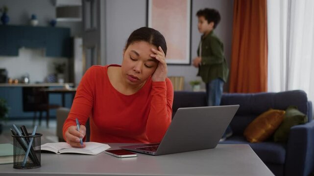 Stress busy African American business woman mother businesswoman freelancer mom female girl using laptop working at home writing notes suffer with noisy active boy child kid son jumping couch playing