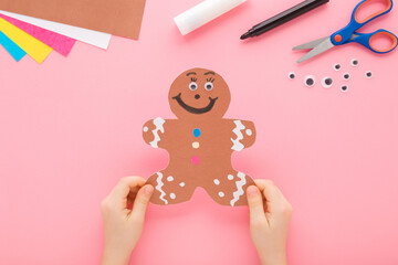 Baby girl hands holding brown big gingerbread man shape from application paper on light pink table background. Pastel color. Little child making decoration. Closeup. Point of view shot. Top down view.