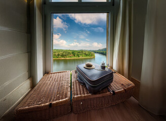 Obraz premium A suitcase sits on a wicker window seat, overlooking a tranquil river and lush green landscape under a vibrant blue sky. The scene evokes a sense of journey and serenity.