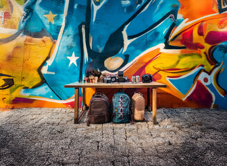 Vibrant graffiti wall backdrop showcases a rustic wooden table displaying various travel bags and photography equipment.  A captivating display of wanderlust and creative expression.