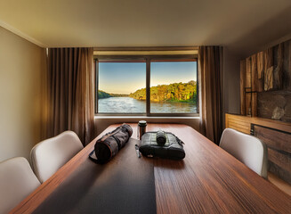 A tranquil river view is framed by a large window in a modern room.  A wooden table with chairs, and exercise items are present, suggesting a space for both relaxation and activity.
