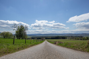 Wide, straight country road surrounded by lush green fields and distant hills under a blue sky with fluffy clouds in Scotland