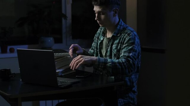 Student while studying for exams fell asleep at night on desk with cup of coffee. Wakes up and continues working on a laptop surrounded by notes. Fatigue and deadlines overwhelm him.