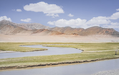 The high-mountain river Murghab flows and meanders among green meadows and fields in a mountain gorge against the background of mountain ranges with rocks and glaciers, near the city of Murgab