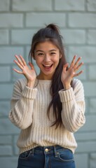 A cheerful young woman with long, dark hair styled in a ponytail beams with joy, her hands raised in a playful gesture. Dressed in a cozy cream sweater against a soft green brick background, she