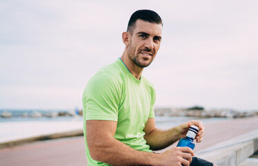 Portrait of muscular male jogger 30 years old looking at camera during workout break at embankment,...