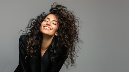 Fototapeta premium An elegant woman with curly, beautiful hair, smiling warmly, dressed in an edgy and fashionable outfit, standing against a clean gray background