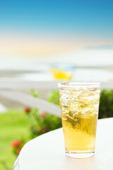 A glass of beer served on a white table, with ice cubes glistening in the natural light and a blurred beach view in the background with vertical copy space. 