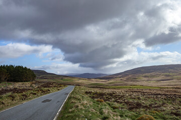 Scenic Scottish countryside road under dramatic clouds with rolling hills in the background during midday