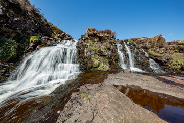 Obraz premium Waterfalls cascading over rocky terrain in the Isle of Skye, Scotland, under a clear blue sky during daytime