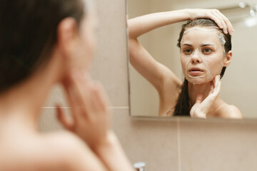Obraz premium Thoughtful young woman applying facial mask in bathroom, looking into mirror, focusing on skin care routine. Self-care and beauty concept.