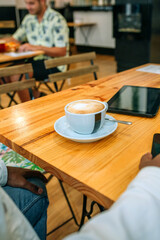 Unrecognizable man sitting in front of cappuccino served in a modern cup and saucer on wooden table of gourmet cafe with tablet and smartphone next to it, while other customers eating on background.