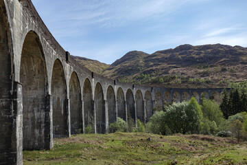 Obraz premium Glenfinnan Viaduct stands majestically against the Scottish Highlands under a clear blue sky during a sunny afternoon