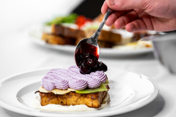 A hand holds a spoon, adding berry sauce to a beautifully decorated layered dessert. The vibrant purple frosting contrasts with the creamy base, showcasing an appealing culinary creation.