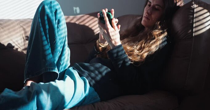 A young woman is lying on the couch at home, casually resting while using her smartphone. She chats and scrolls through newsfeeds, enjoying some downtime. Home relaxation and social media concept.