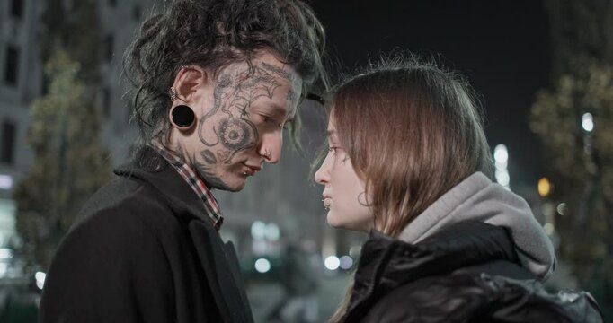An informal couple, a guy and a girl with dreadlocks, tattoos and facial piercings in casual clothes, hitchhike on the street of a night city and touch foreheads while standing close to each other