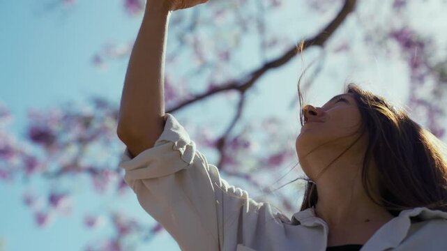 Close-up of a young woman enjoys a warm day in a park, taking pictures with her phone of vibrant flowers of a jacaranda tree under a clear blue sky