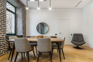 Interior with a dining table in the loft-style apartment. There is a large leather .armchair in the corner of the room. Brick wall with a window.