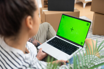 laptop with an empty green mock-up screen, held amidst cardboard boxes, signifies a young couple's...