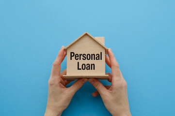 A wooden house model with Personal Loan text displayed, held against a blue background, symbolizing...