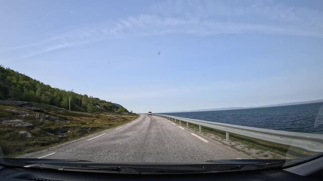 Driving along Norway's northern tundra on E69 highway. Rocks and low trees line the left side, while the sea stretches along the right. A serene and picturesque coastal journey.