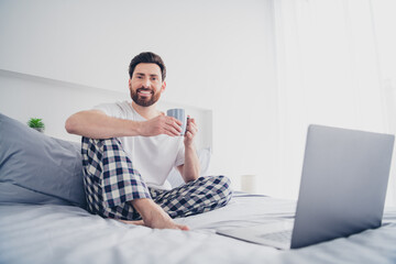 Photo of cheerful positive young man dressed nightwear lying bed drinking coffee watching modern...