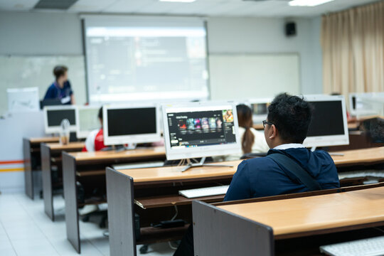 Students are seated at desks using desktop computers in a classroom, focusing on their tasks in a learning environment equipped with technology.