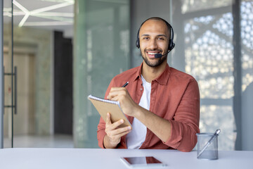 Webcam view, a man with a headset and a notepad in his hands looks into the camera, records data while listening to an online meeting and conference with colleagues and partners.