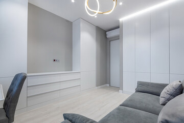 A room in an apartment with a modern design. White room decor, built-in wardrobe with TV space, gray sofa with pillows.