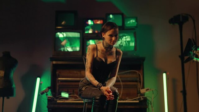 Depressed offbeat young woman sitting alone in strange studio or basement. Static portrait of frustrated freaky lady with tattooed body and piercing on face, abandoned and misunderstood by society