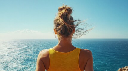 Elegant woman runner in yellow top gazing at tranquil blue sea from cliff edge, enjoying calming ocean breeze on sunny day.